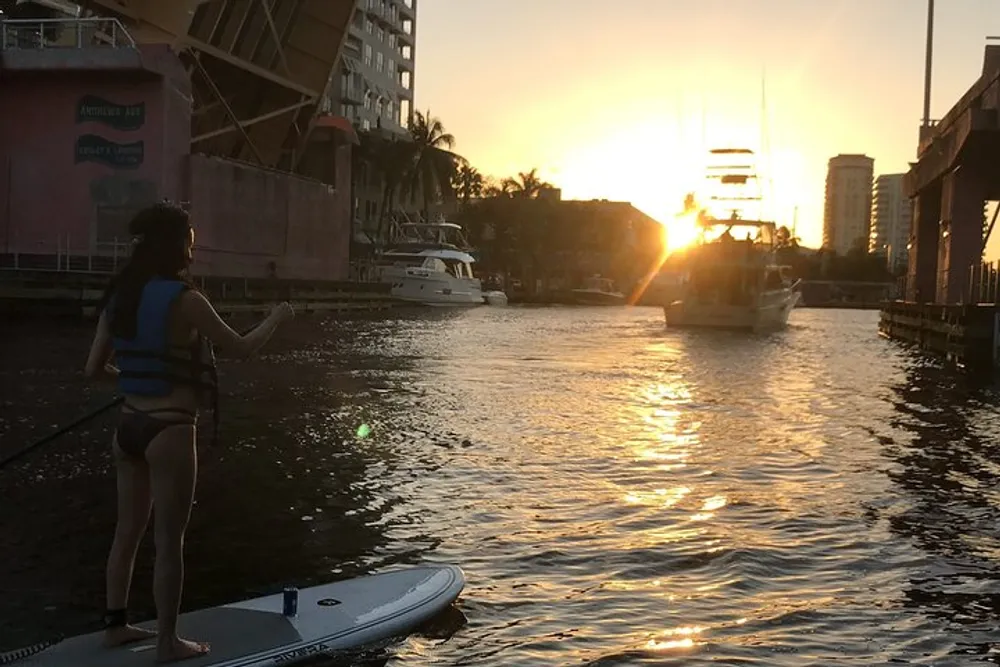 A person is paddleboarding in a calm waterway during a beautiful sunset with boats and city buildings in the background