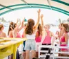 A group of people wearing shades of pink and white is enjoying a lively gathering on a boat with some raising their arms in what appears to be a cheerful celebration