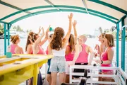 A group of people wearing shades of pink and white is enjoying a lively gathering on a boat, with some raising their arms in what appears to be a cheerful celebration.