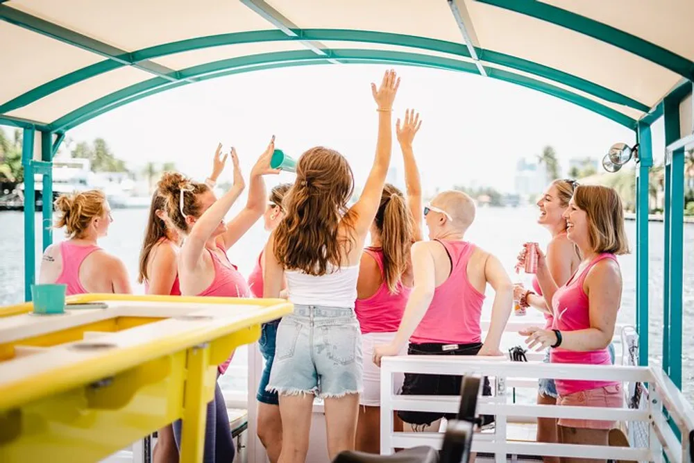 A group of people wearing shades of pink and white is enjoying a lively gathering on a boat with some raising their arms in what appears to be a cheerful celebration