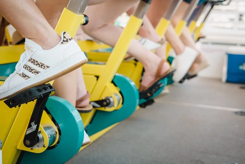 The image shows a row of individuals on indoor cycling machines focusing on their feet which are strapped into the pedals