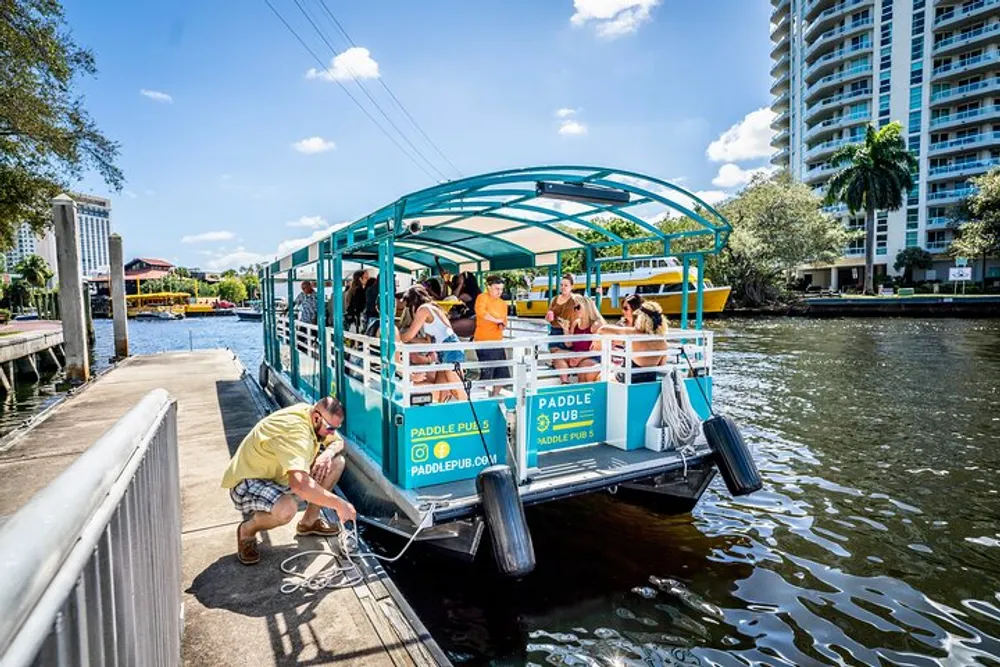 A group of people is enjoying a sunny day on a pedal-powered boat bar along a river with a person mooring the boat to the dock