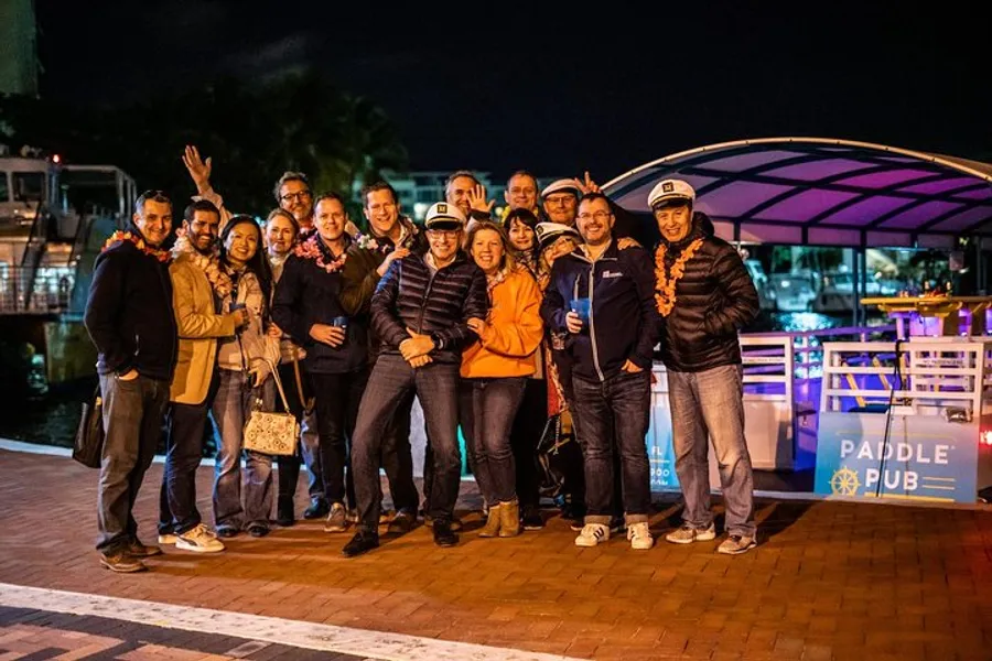 A group of smiling people is posing for a photo at night in front of a boat with a sign saying PADDLE PUB.