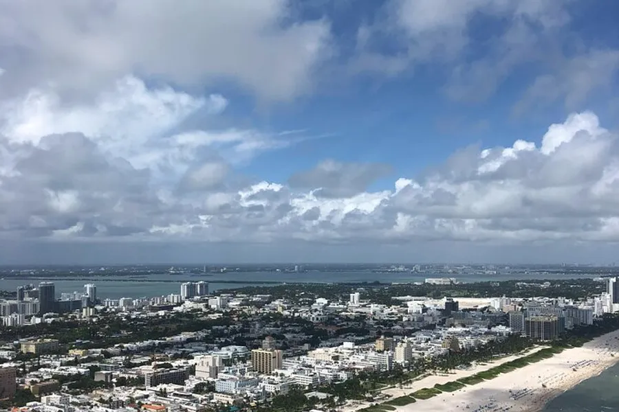 This image features an expansive aerial view of a coastal city with numerous buildings adjacent to a beach, under a sky dotted with cumulus clouds.