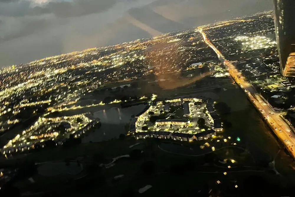 The image shows a nighttime aerial view of a city with glowing streets and buildings taken from an airplane with a sense of motion blur