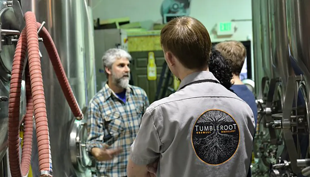 People are engaged in a discussion or tour inside a brewery called Tumbleroot as indicated by the logo on the back of a persons shirt