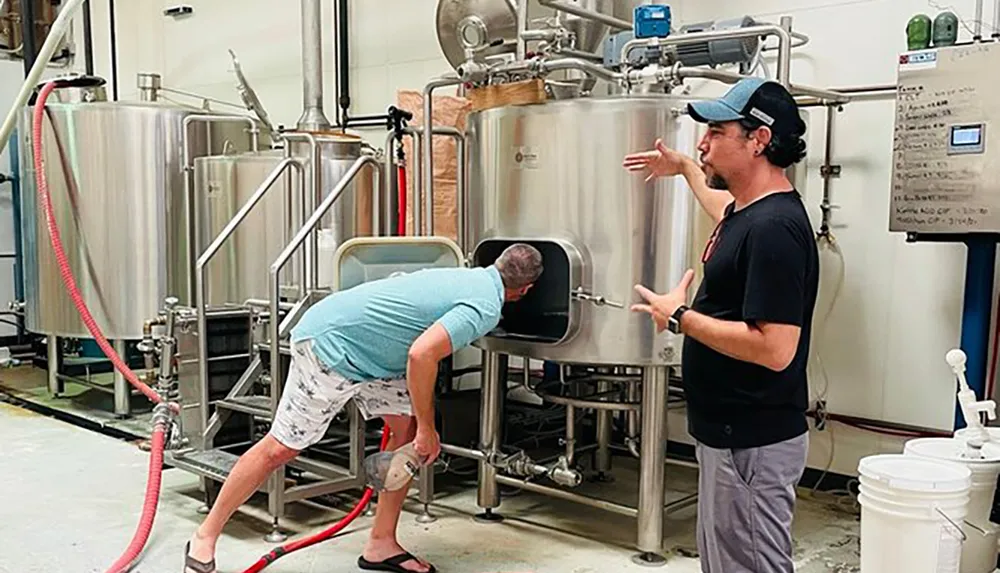 One person is looking inside a large brewing tank while another person gestures and possibly explains the brewing process in a brewery