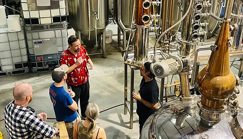 A group of people is taking a tour of a distillery listening to an individual who appears to be an employee explaining the process while standing amongst stainless steel tanks and copper stills