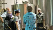 A group of people is touring a brewery, learning about the beer production process among stainless steel fermentation tanks.
