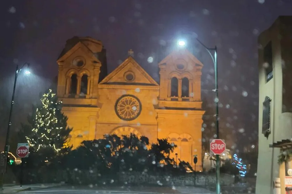 An illuminated church stands prominently at night with a decorated Christmas tree nearby viewed through a rain-spattered window