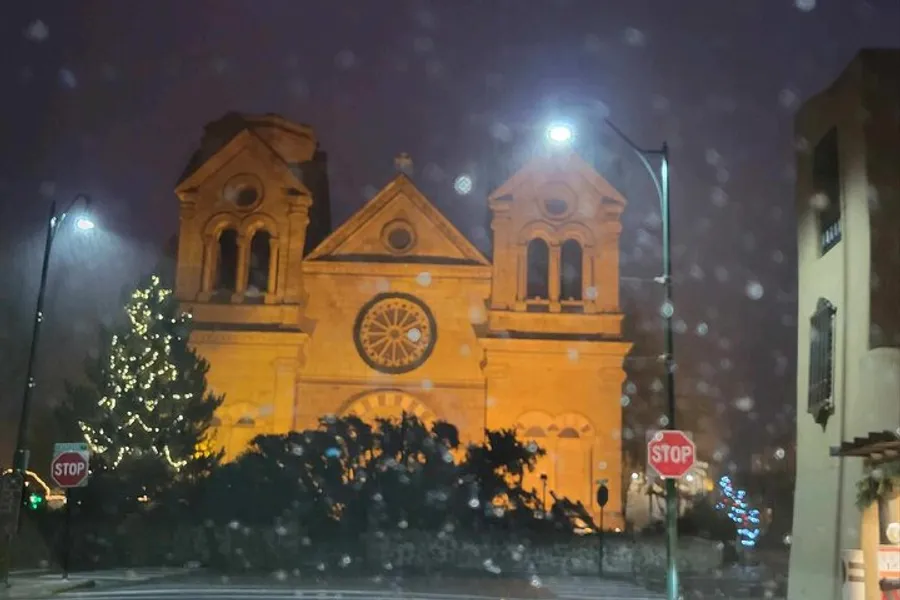 An illuminated church stands prominently at night with a decorated Christmas tree nearby, viewed through a rain-spattered window.
