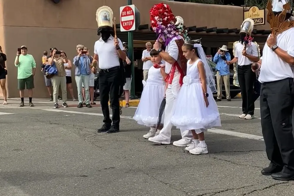 A procession with two girls in white dresses and a person wearing a mask with a flower headdress is taking place on a street while spectators watch from the sidelines