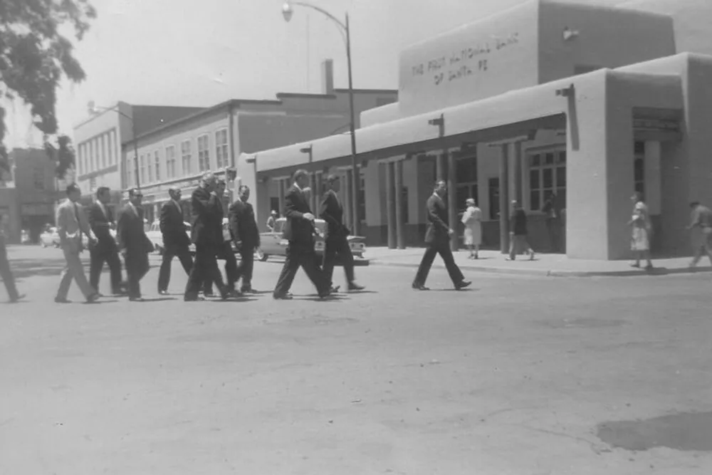 The image shows a black-and-white photo of a group of people likely men in business attire crossing a street in front of what appears to be a mid-20th century American downtown with a clear emphasis on the historical context suggested by the car models and architectural styles visible