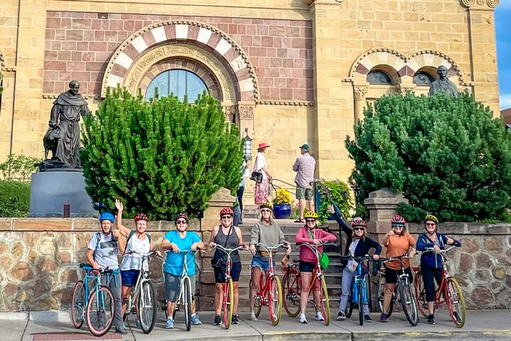 A group of people wearing helmets poses with their bicycles in front of a historic building and a statue