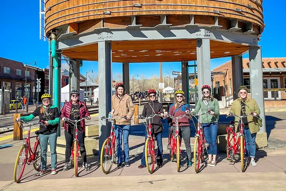 A group of smiling people wearing helmets are standing with bicycles under a wooden structure on a sunny day