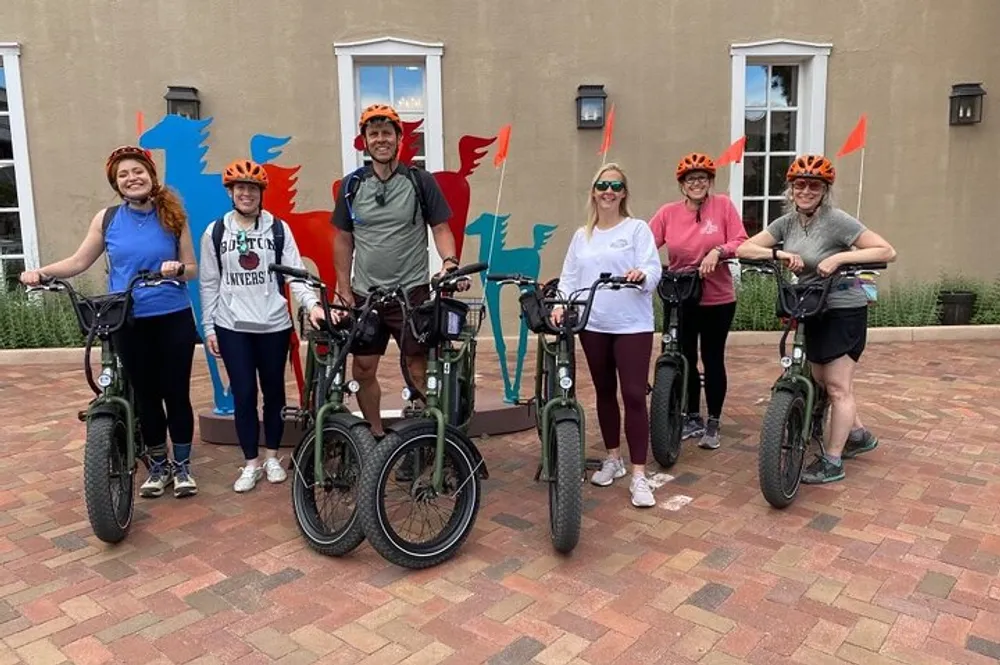 A group of people wearing matching orange flame-themed helmets are posing with their bicycles smiling for a photo in a courtyard