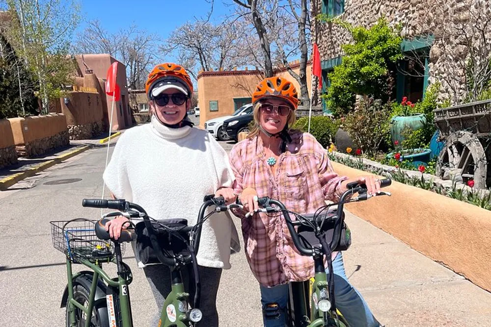 Two individuals wearing orange helmets are standing with their bicycles on a sunny day smiling for the camera on a street with cars and greenery in the background