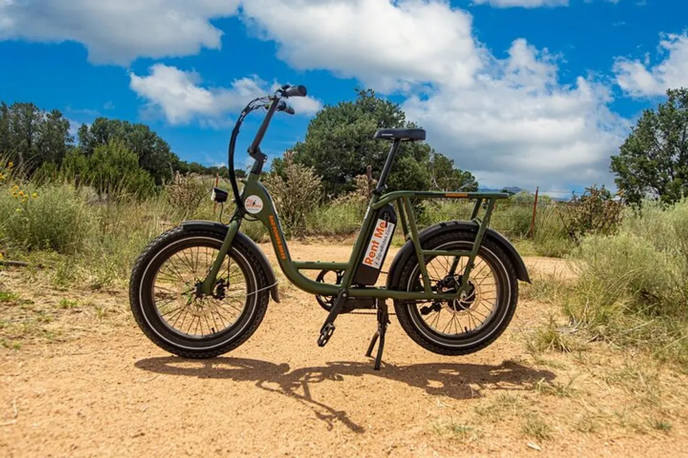 An electric bicycle is parked on a dirt path with a backdrop of blue sky and scattered clouds surrounded by brush and trees