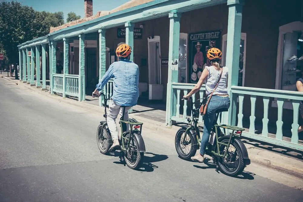 Two people wearing helmets are riding bicycles past a row of vintage-looking storefronts on a sunny day