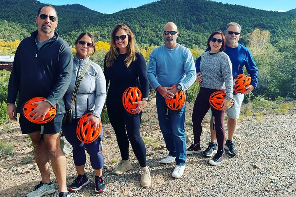 A group of six adults are standing outdoors holding orange helmets posing for a photo with a backdrop of trees and hills