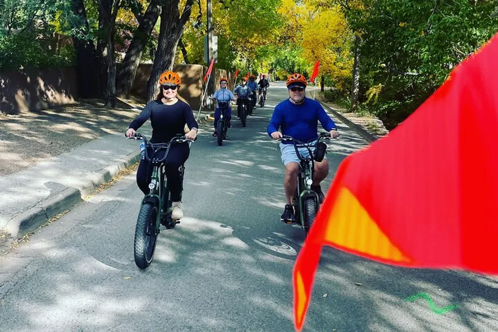 A group of people are enjoying a bicycle ride on a tree-lined street with one person in the foreground somewhat obscured by a red flag in motion