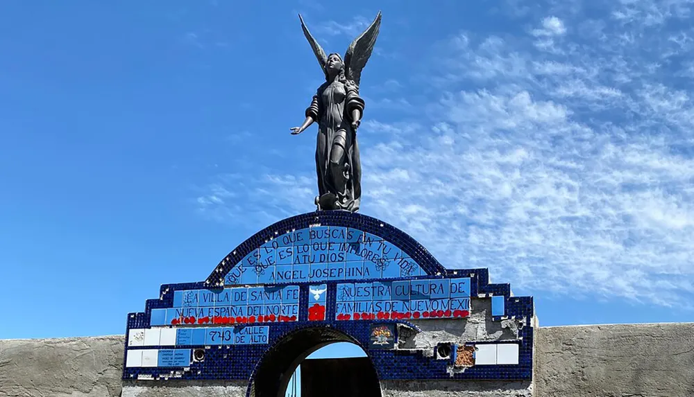 The image shows a statue of an angel perched atop an arch with blue mosaic and text against a backdrop of a strikingly blue cloud-flecked sky