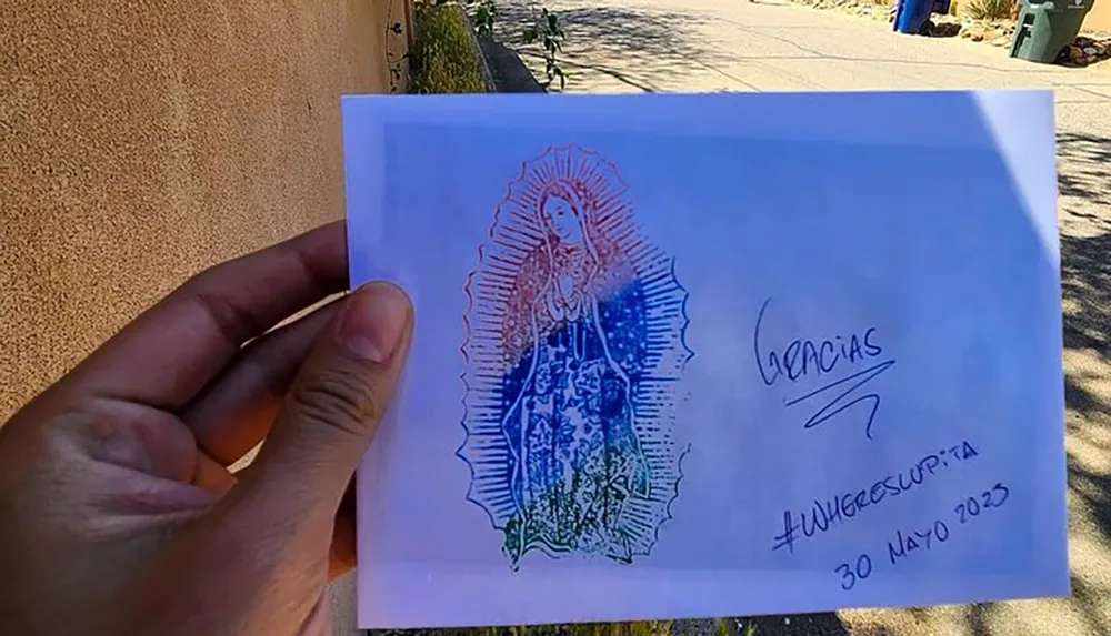 A persons hand is holding a piece of paper with a printed image reminiscent of the Virgin Mary and inscriptions that include Gracias a hashtag and a date 30 Mayo 2023