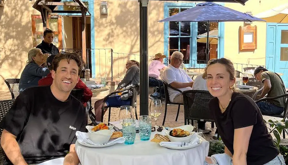 Two people are smiling at the camera while sitting at a table with food and drinks on a sunny outdoor patio at a restaurant