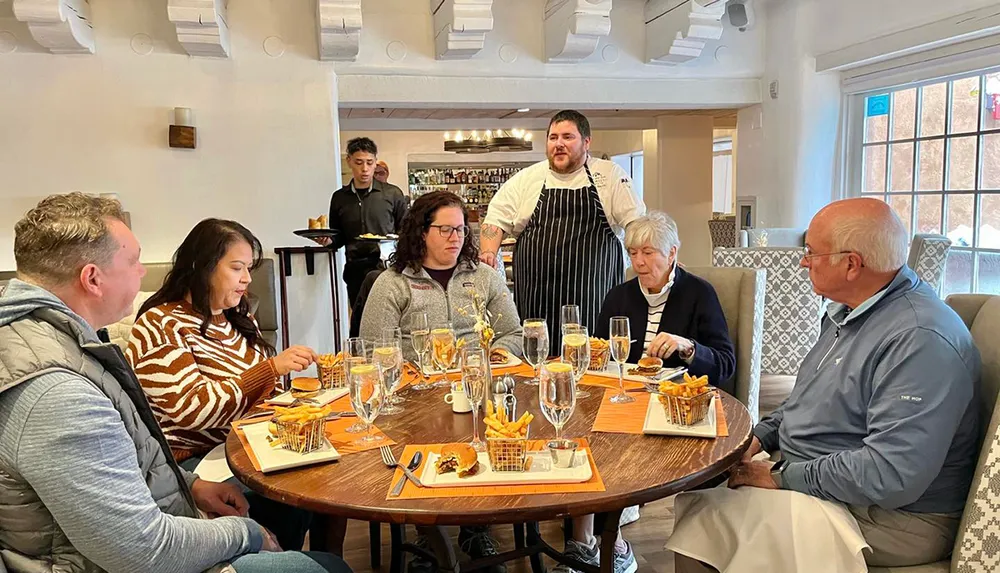 A group of people are dining at a table with a chef standing behind them in what appears to be a cozy restaurant setting