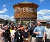 A group of smiling individuals is posing for a photo in front of a wooden water tank marked Santa Fe Railyard under a clear blue sky