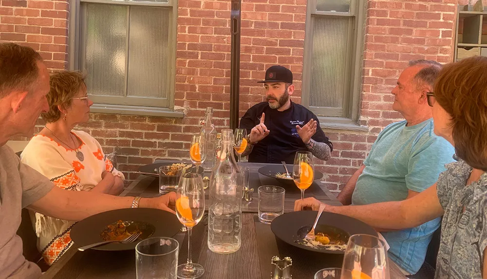 A group of people are enjoying a meal outdoors engaged in conversation with a man who appears to be explaining something with expressive hand gestures