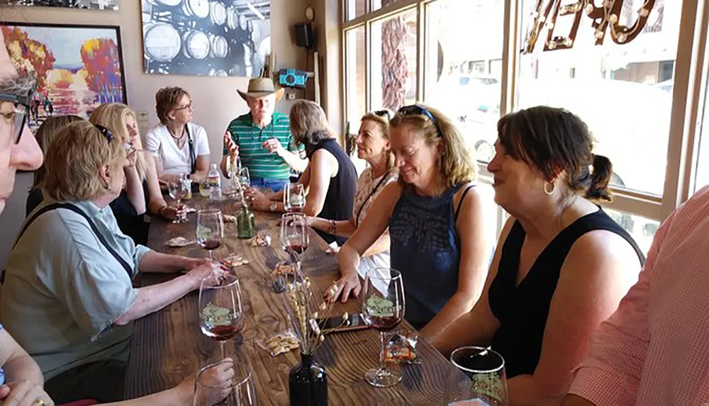 A group of people appears to be enjoying a wine tasting event at a long table inside a room with artwork on the walls