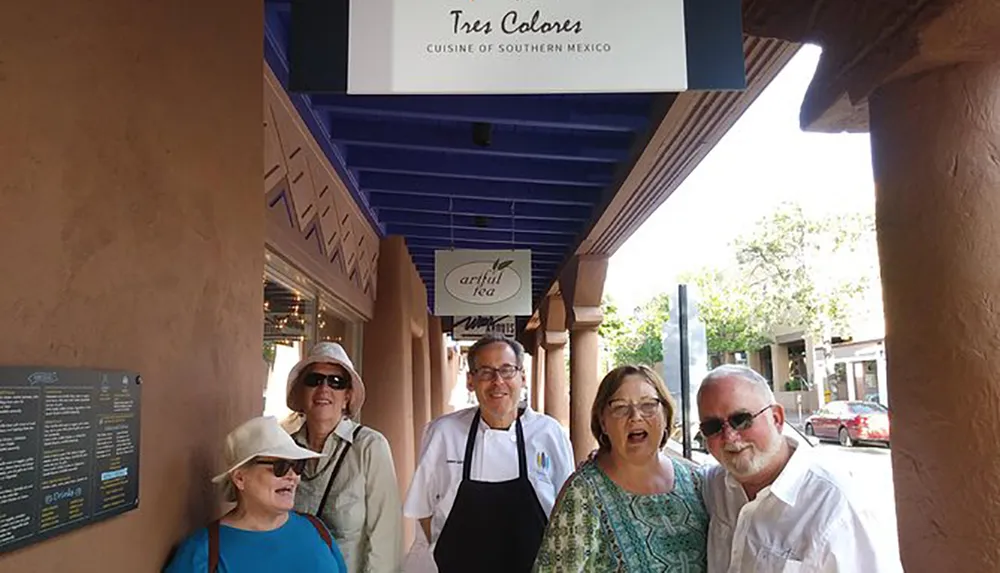 A group of five people is smiling and posing for a photo under a restaurant sign that reads Tres Colores CUISINE OF SOUTHERN MEXICO