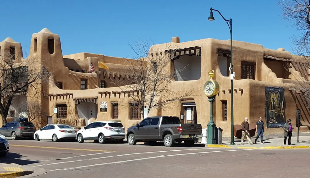 This image shows a street view with people walking near traditional adobe architecture cars parked on the side and a clear blue sky