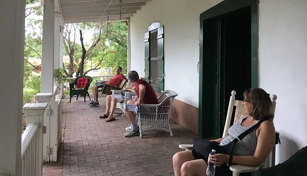 Three people are relaxing on rocking chairs on a shaded porch with a brick floor surrounded by a natural setting