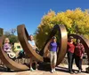 A group of people is posing and smiling in front of a large abstract metal sculpture under a clear blue sky with a backdrop of autumn-colored trees
