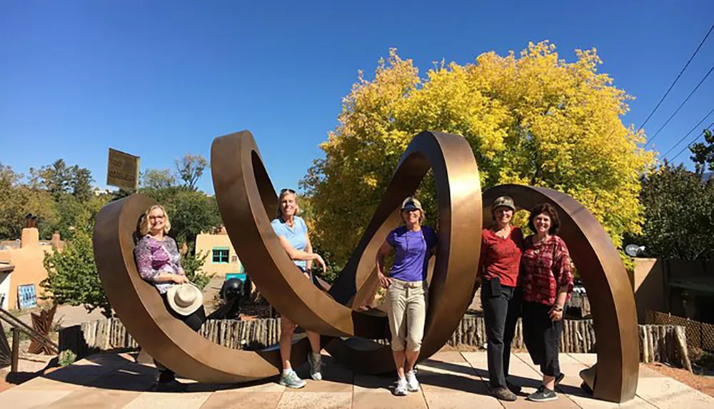 A group of people is posing and smiling in front of a large abstract metal sculpture under a clear blue sky with a backdrop of autumn-colored trees