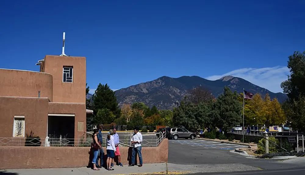The image features a small group of people engaged in conversation in front of a building with Pueblo-style architecture with a mountain range and a clear blue sky in the background