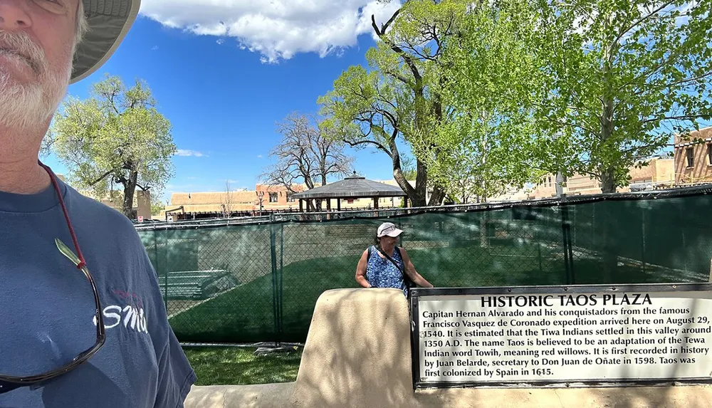 The image shows a partial selfie of a person with a sign that reads Historic Taos Plaza along with another person seated on a bench against a backdrop of greenery and buildings