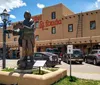 A statue of a person playing a guitar stands in front of the Hotel La Fonda under a clear blue sky surrounded by parked cars and typical Southwestern architecture