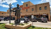 A statue of a person playing a guitar stands in front of the Hotel La Fonda under a clear blue sky, surrounded by parked cars and typical Southwestern architecture.