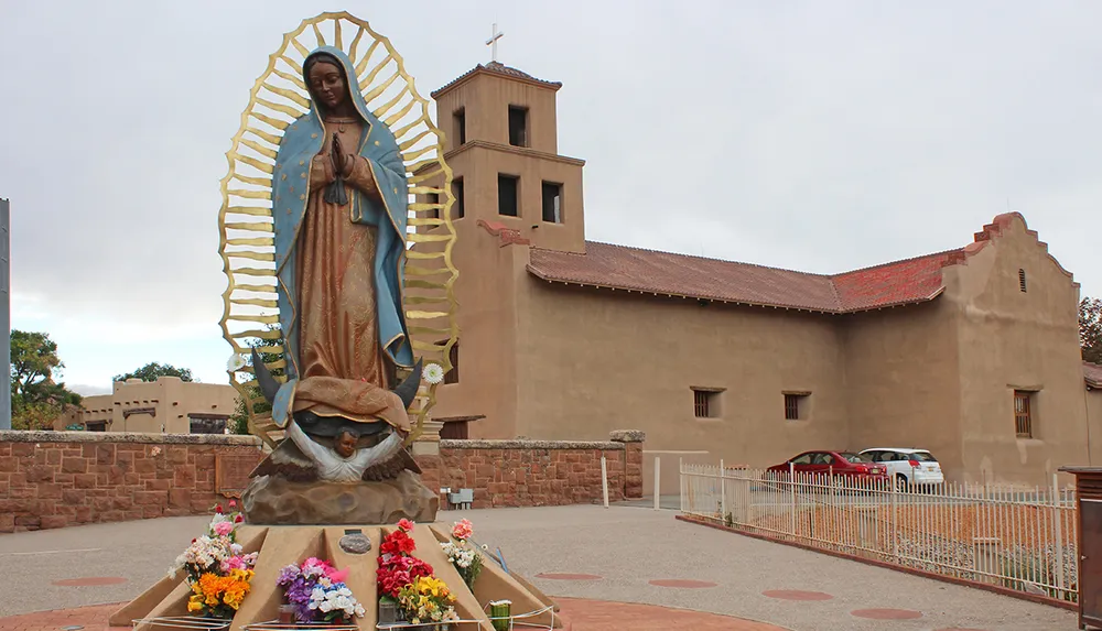 The image features a large colorful statue of the Virgin Mary in front of an adobe-style church building adorned with flowers at its base