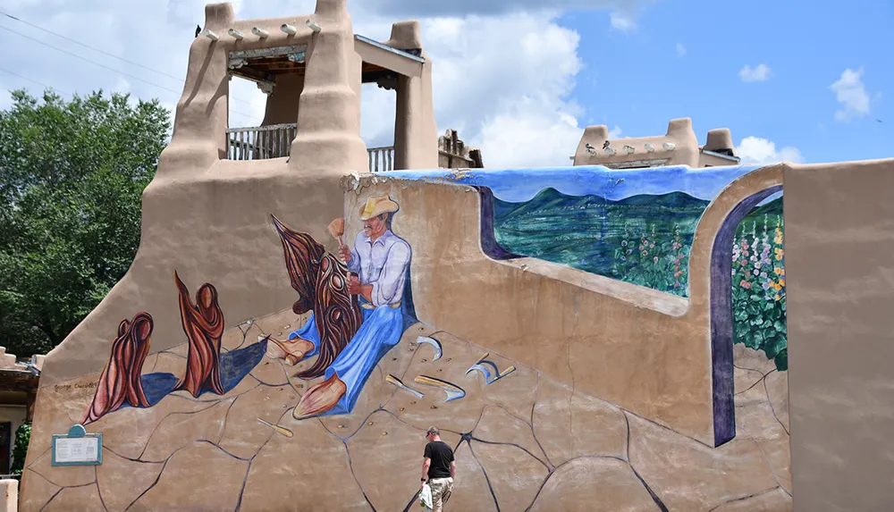 This image shows a person walking past a large colorful mural painted on the side of an adobe building depicting an artist at work with a landscape scene and floral patterns