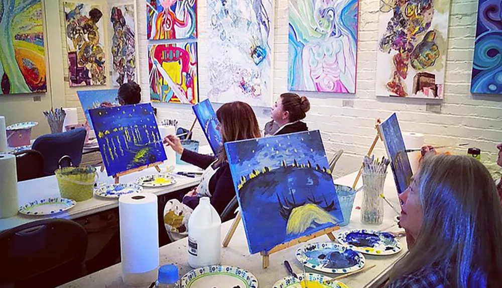 A group of people are attending an art class or workshop painting on canvases in a studio surrounded by colorful artwork on the walls