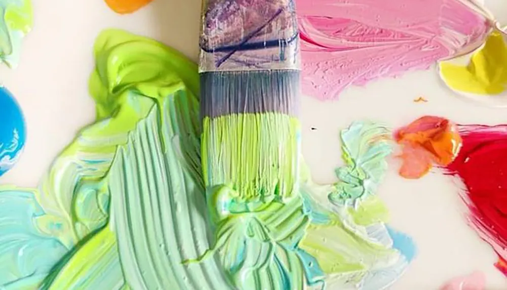 A paintbrush is dipped into thick brightly colored paints spread on a palette