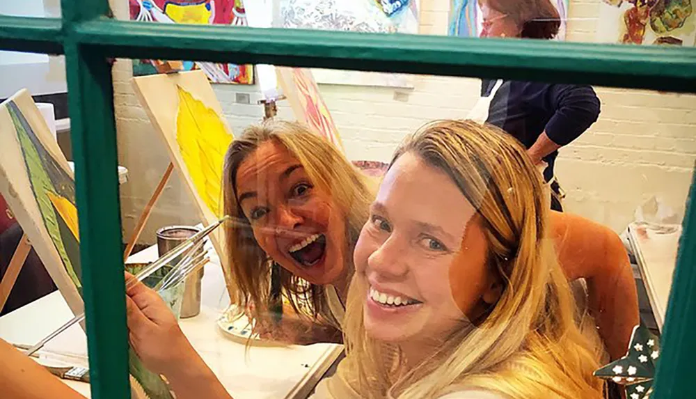 Two joyful women are peeking through a green window frame at a painting class