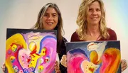 Two people are holding vibrant, abstract paintings with heart shapes as the central theme.