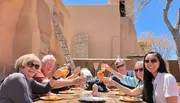 A group of smiling people are toasting with drinks at an outdoor dining table under a clear blue sky.