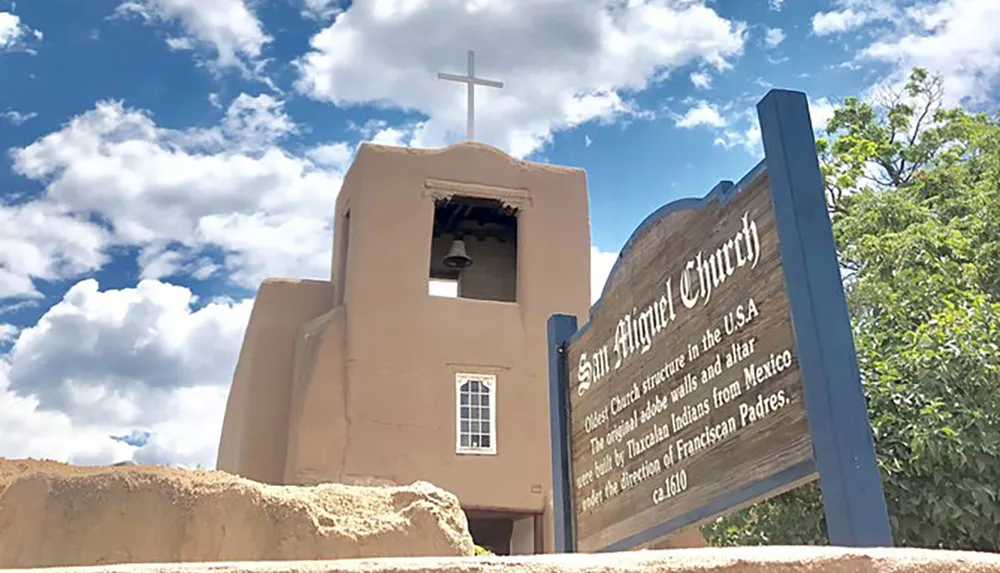 The image shows the San Miguel Church described on the sign as the Oldest Church structure in the USA with its adobe walls and altar dating back to the time of the Tlaxcala Indians from Mexico and Franciscan Padres circa 1610 set against a blue sky with scattered clouds