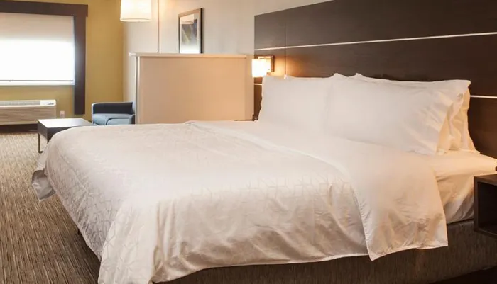 The image displays a neatly made hotel room bed with white linens in a modernly furnished room with a seating area near the window